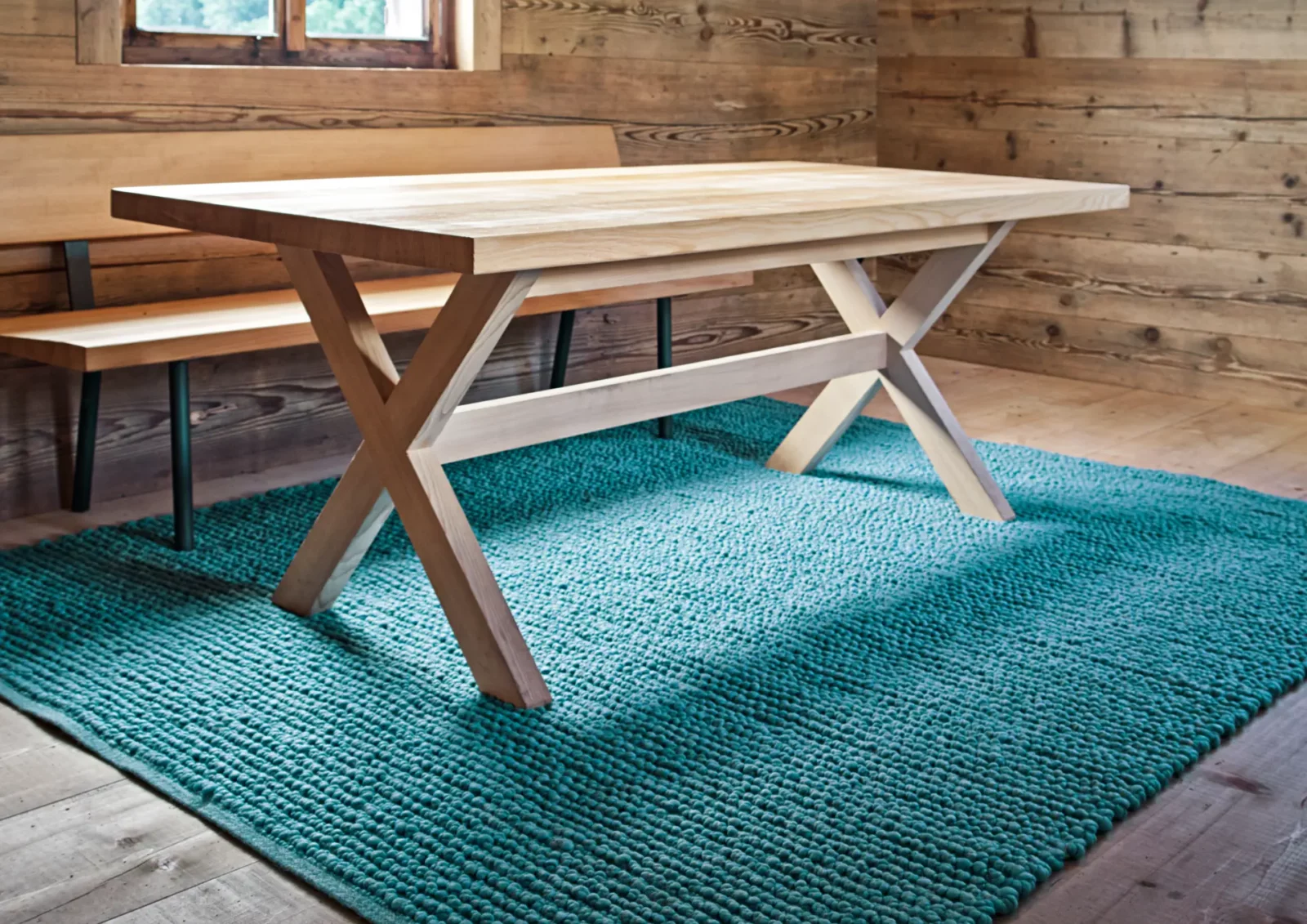 Holztisch mit X-förmigen Beinen auf türkisfarbenem Teppich.