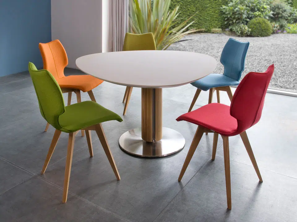 Runder Tisch mit Holzfuß, umgeben von bunten Stoffstühlen in Grün, Blau, Rot, Gelb und Orange.